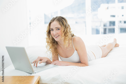 Pretty cheerful blonde lying in bed using laptop