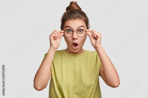 Horizontal portrait of stunned lovely young woman stares with jaw dropped, looks in bewilderment through round spectacles, can`t believe in shocking news, poses against white studio wall. Emotions