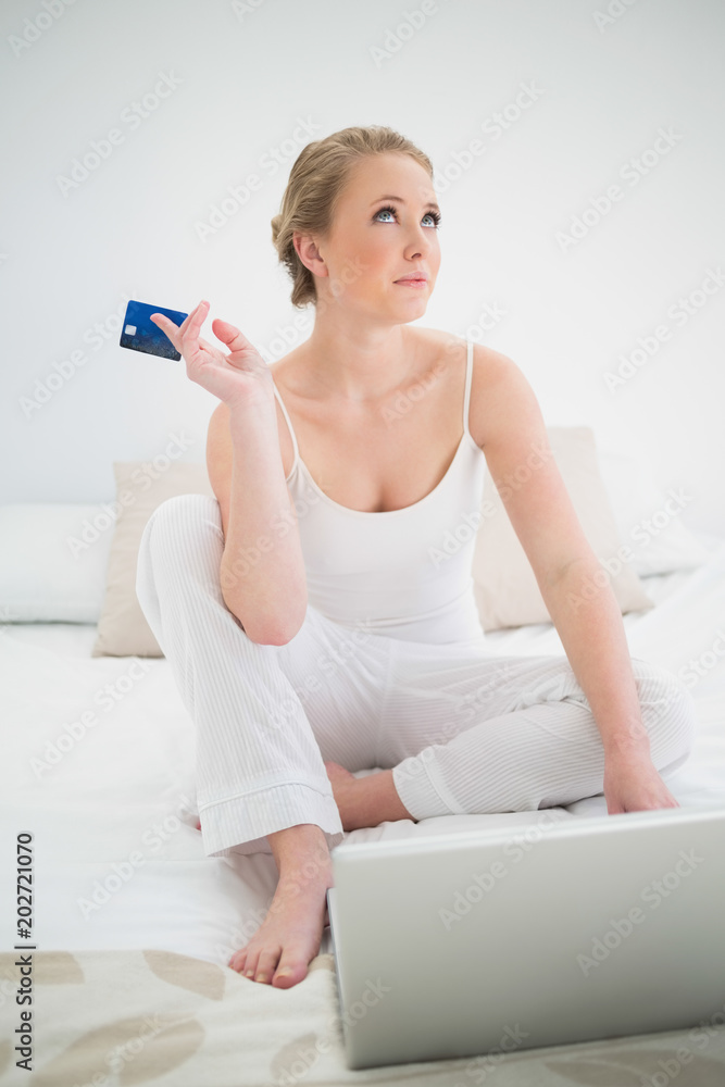 Natural thoughtful blonde holding credit card and sitting in front of laptop Natural thoughtful blonde holding credit card and sitting in front of laptop