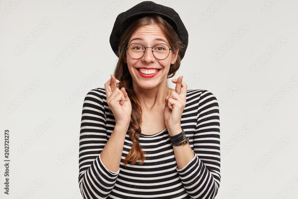 Attractive young female Parisian, has French style of clothing, happy expression, crosses fingers, wishes to have trip around world, isolated over white background. Pretty woman in stylish beret Attractive young female Parisian, has French style of clothing, happy expression, crosses fingers, wishes to have trip around world, isolated over white background. Pretty woman in stylish beret