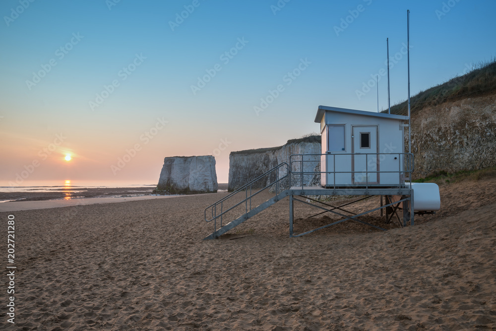 Lifeguard hut on empty beach during colorful sunrise with rock cliffs in background