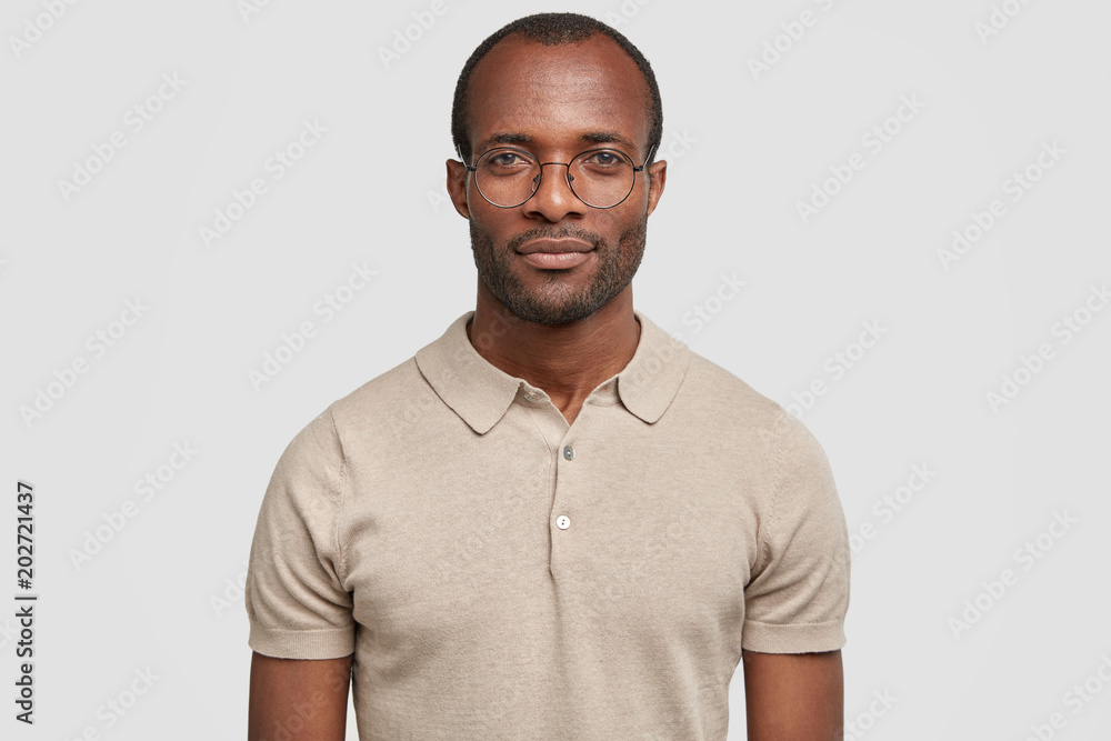 Fototapeta premium Confident young male manager with dark healthy skin, wears round spectacles and casual t shirt, looks seriously at camera, being sure in everything, has stubble, poses against white background