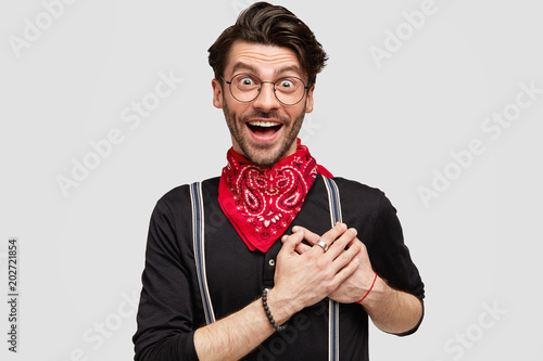 Cheerful overjoyed hipster with dark stubble and trendy hairdo, exppresses his amazement and thankfulness, keeps hand on heart, has excited expression, poses indoor. People, human emotions concept