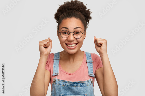Studio shot of lovely joyful African American female clenches fists with great success and positiveness, has broad smile and white even teeth, isolated over studio background. Happiness and victory