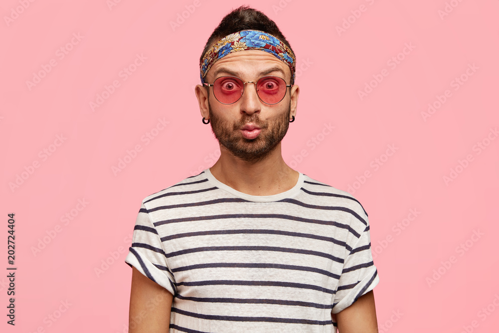 Hippy young fashionable male model with stylish sunglasses, headband, earrings and striped t shirt, being stunned to recieve shocking news, demonstrates freedom, isolated over pink studio background Hippy young fashionable male model with stylish sunglasses, headband, earrings and striped t shirt, being stunned to recieve shocking news, demonstrates freedom, isolated over pink studio background