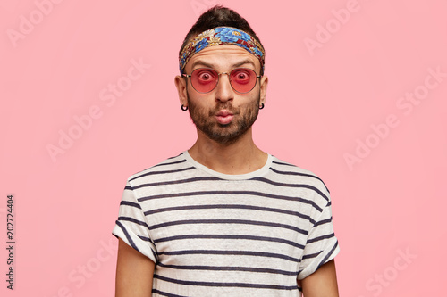 Hippy young fashionable male model with stylish sunglasses, headband, earrings and striped t shirt, being stunned to recieve shocking news, demonstrates freedom, isolated over pink studio background