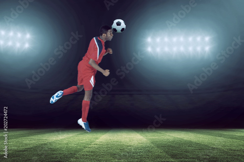 Football player in red jumping against football pitch under spotlights © vectorfusionart