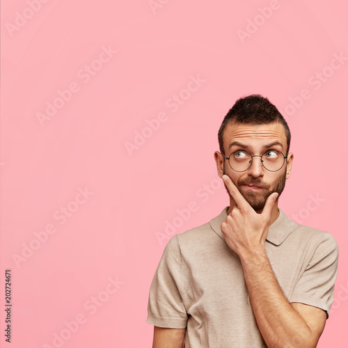 Vertical shot of pensive bearded young male holds chin and contemplates about something, wears round spectacles and t shirt, poses against pink background with copy space for your advertisement