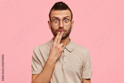 Attractive young male model with amazed shocked expression, looks with widely opened eyes att camera, has stubble, wears casual t shirt, isolated over pink background. Caucasian unshaven man