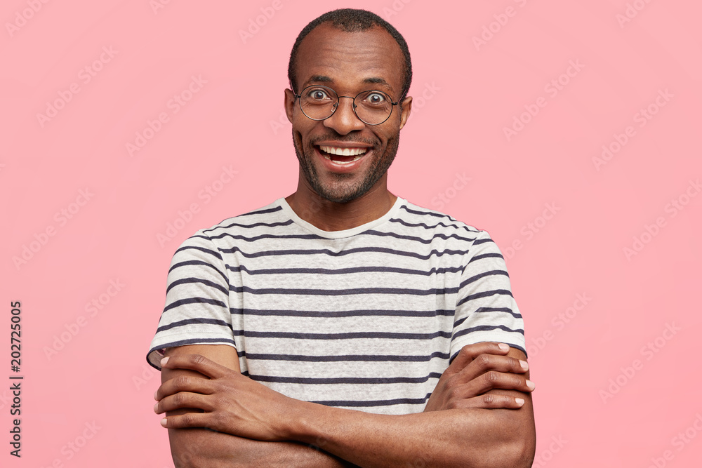 Pleased African American male with positive smile, keeps arms folded, being glad to recieve good comments of his work. Happy dark skinned student rejoices successfully passed exam, isolated on pink Pleased African American male with positive smile, keeps arms folded, being glad to recieve good comments of his work. Happy dark skinned student rejoices successfully passed exam, isolated on pink