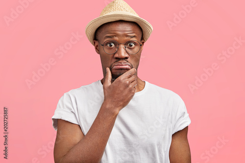 Hesitant dark skinned male keeps chin and looks with stunned expression at camera, can`t understand something, curves lips in bewilderment, isolated over pink background. Surprisement and people