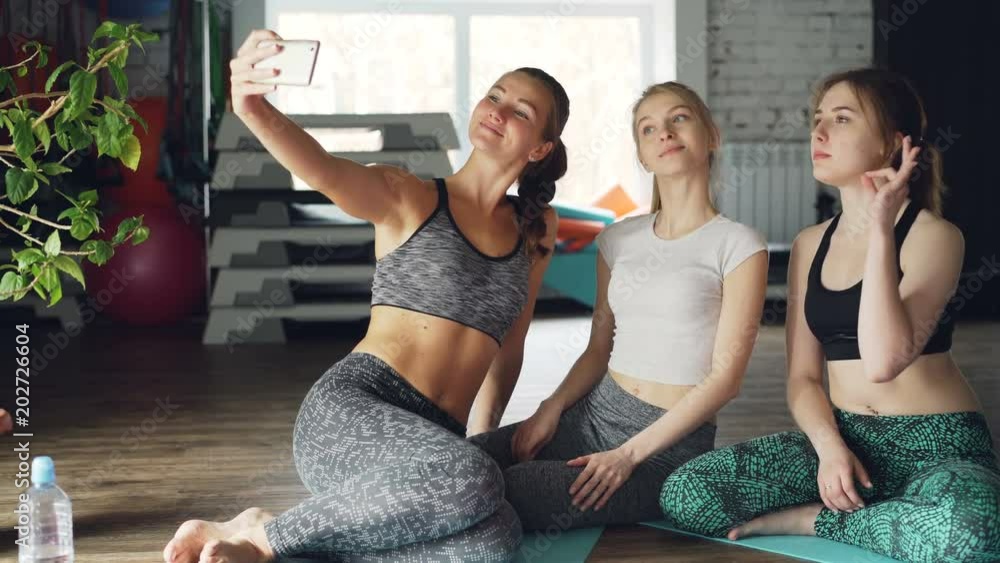 Attractive young women are taking selfie after yoga class in light modern gym in wellness center. Women are wearing trendy sports clothes. Sports equipment in background. Attractive young women are taking selfie after yoga class in light modern gym in wellness center. Women are wearing trendy sports clothes. Sports equipment in background.