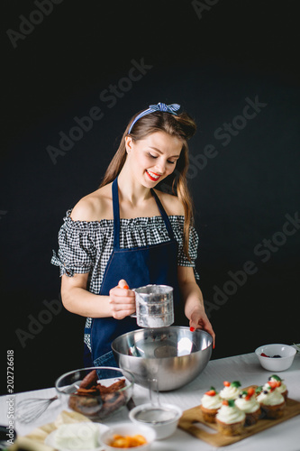 Pastry woman sifting white flour to the bowl, freshly made cupcakes on the table near photo