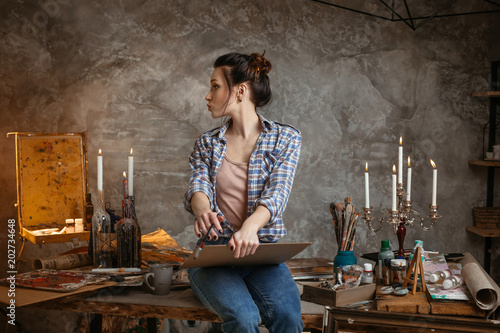 Woman painter sitting on the table, drawing and looking left. Creative concept. Drawing supplies, oil paints, artist brushes, canvas, candle.