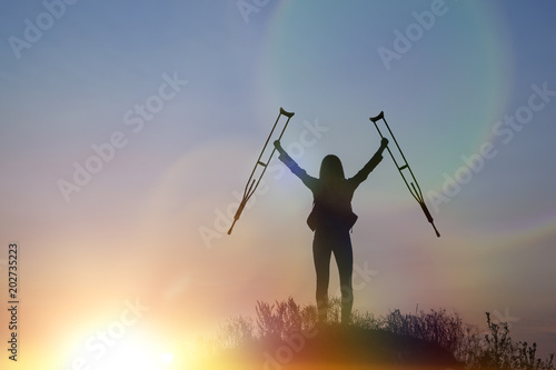 silhouette of a happy girl on top of a mountain with crutches, lifting her arms up, facing dramatic sky at dawn. Defeat of the disease, rehabilitation, healing, trauma, medicine. Prove what you can do