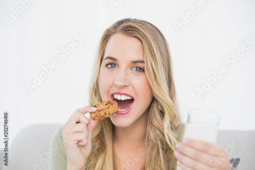 Cheerful cute blonde eating cookie with milk sitting on cosy sofa