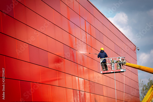 worker of Professional Facade Cleaning Services washing the red wall. Worker wearing safety harness washes wall facade at height on modern building in a crane. photo