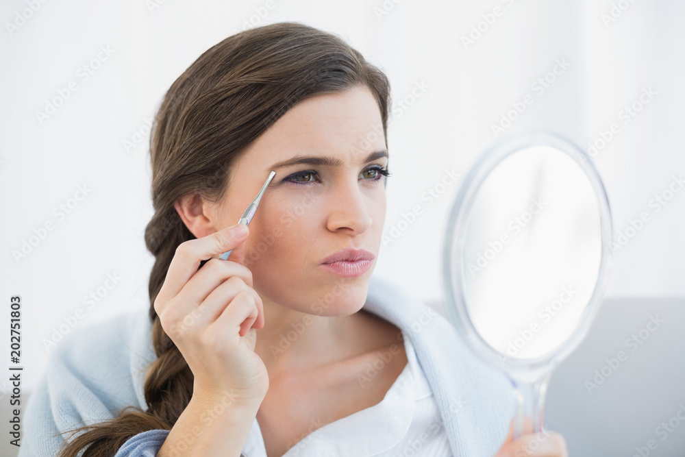 Concentrated casual brown haired woman in white pajamas plucking her eyebrows Concentrated casual brown haired woman in white pajamas plucking her eyebrows