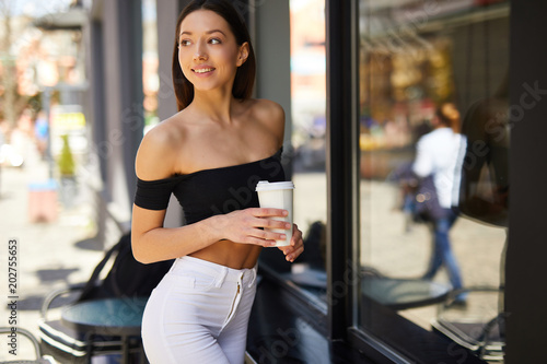 Brunette girl stand on the street look at camera and dring her delicious coffee from white cup, spring time photo shoot