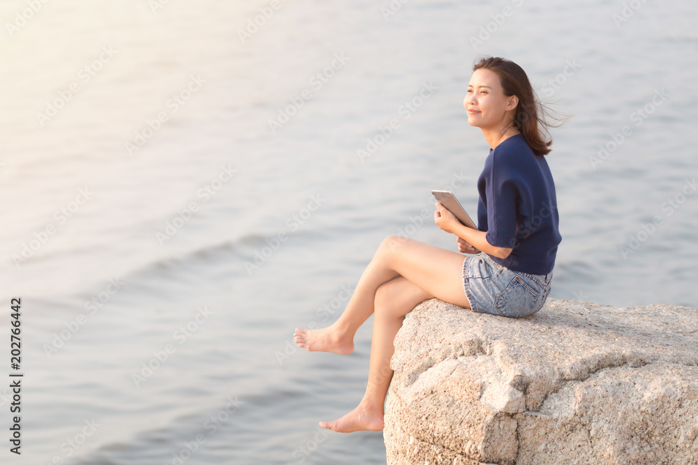 Happiness young Asian woman relaxing and sitting on the rock cliff with smiling face and think about something good while holding or using digital tablet with blurred sea wave ocean background.