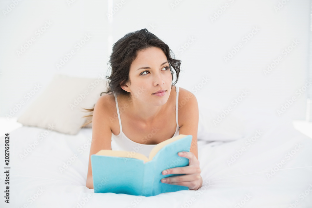 Thoughtful casual brunette in white pajamas relaxing on a bed reading a book Thoughtful casual brunette in white pajamas relaxing on a bed reading a book