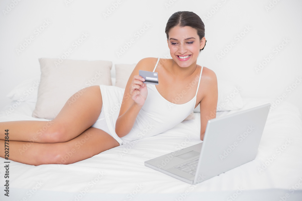Happy young brown haired model in white pajamas shopping online with her laptop Happy young brown haired model in white pajamas shopping online with her laptop