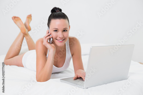 Smiling casual woman using cellphone and laptop in bed