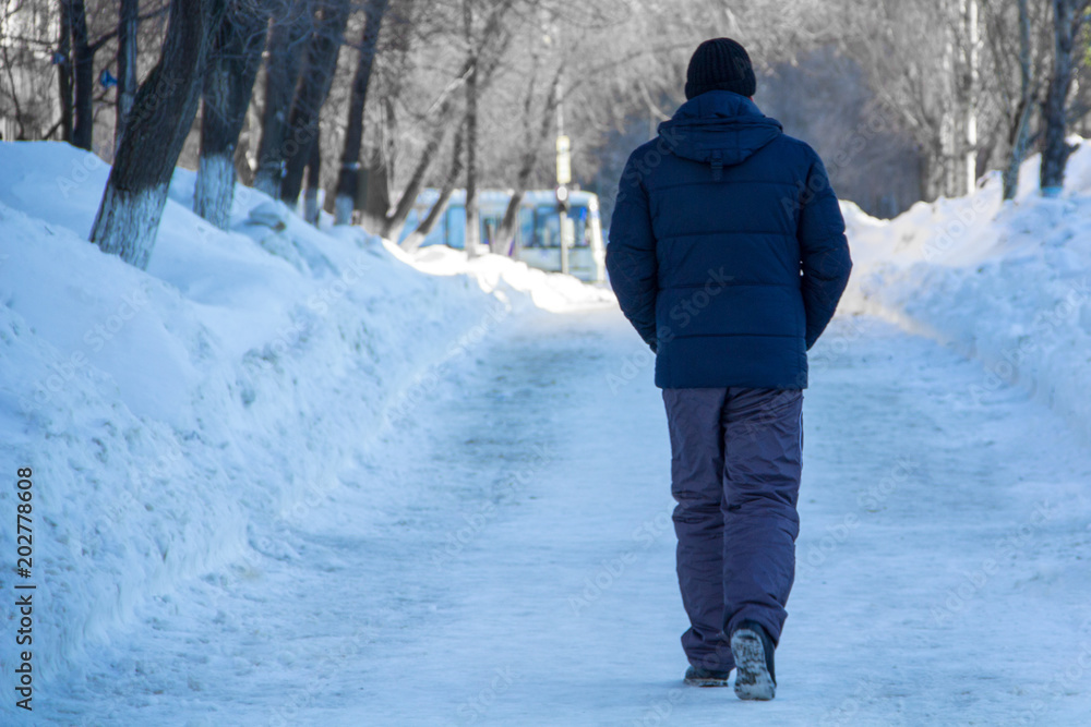 A man in a winter jacket, hat and pants walks along a pedestrian path between snowdrifts and trees across the city.