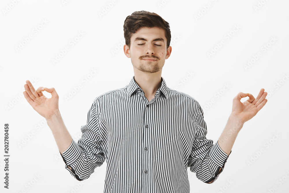 Naklejka premium Portrait of charming satisfied caucasian man with relaxed expression, spread hands in zen gesture, standing with closed eyes and pleased calm smile, meditating or practicing yoga over gray wall