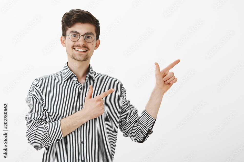 Studio shot of positive charming friendly man with moustache and beard in stylish round glasses, pointing at upper right corner with finger gun gestures and smiling joyfully, being in great mood Studio shot of positive charming friendly man with moustache and beard in stylish round glasses, pointing at upper right corner with finger gun gestures and smiling joyfully, being in great mood