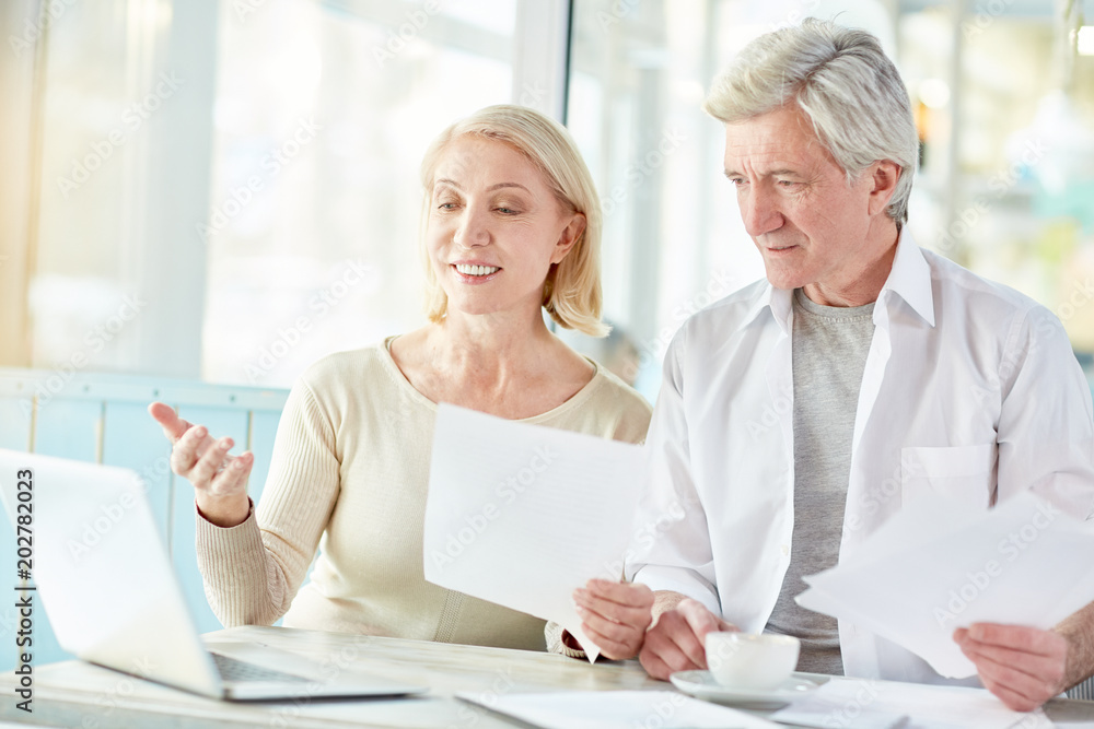 Successful mature businesswoman with papers pointing at laptop display while analyzing online data with colleague Successful mature businesswoman with papers pointing at laptop display while analyzing online data with colleague