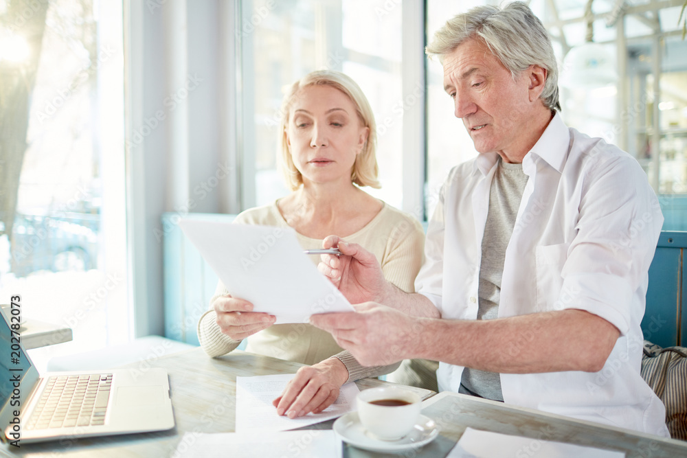 Grey-haired businessman showing his client or colleague some papers to sign at meeting in cafe Grey-haired businessman showing his client or colleague some papers to sign at meeting in cafe