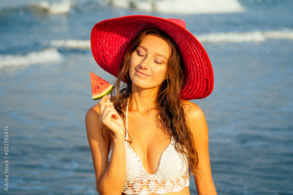 beautiful and young girl in a red straw hat holds a piece of watermelon and enjoys a vacation on the beach. red juicy watermelon on a hot summer day refreshing