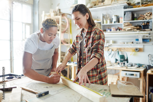 Photos Two young intercultural women measuring length of wooden plank before sawing it
