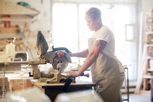 Young female carpenter standing by workbench with electric sawing instrument and using it during work