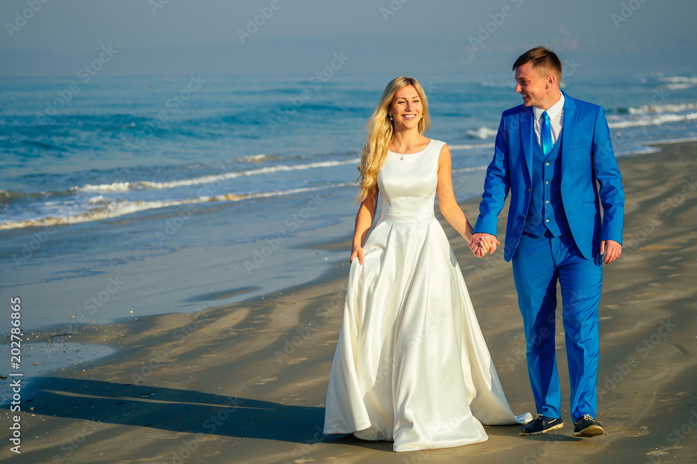 handsome groom in a chic suit and a beautiful bride in a wedding gown walk along the beach. concept of a chic and rich wedding ceremony on the beach handsome groom in a chic suit and a beautiful bride in a wedding gown walk along the beach. concept of a chic and rich wedding ceremony on the beach