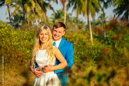 Handsome groom in a luxury suit and a beautiful bride in a beautiful wedding dress smiling and posing on a background of palm trees. concept of a chic and rich wedding ceremony on the beach