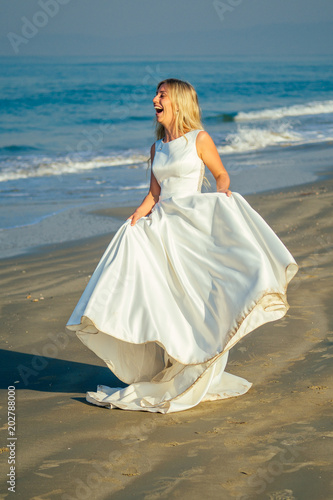 young and beautiful bride with long blond hair smiling and walking on the ocean background in a beautiful white long wedding dress. a happy bride married a wedding ceremony on the beach by the sea