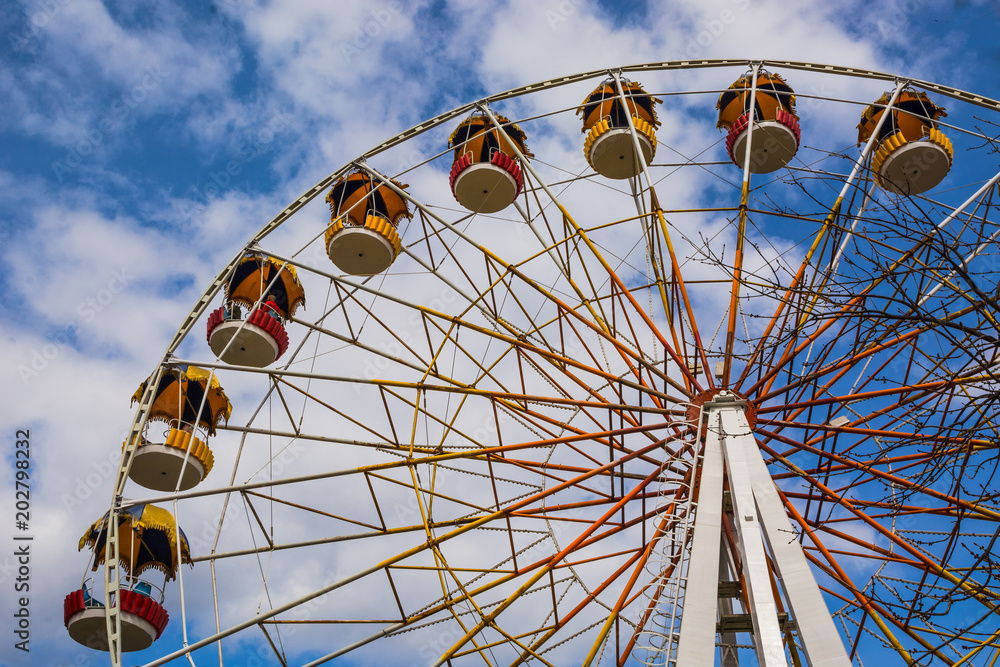 Attraction for entertainment in the city recreation park. A large wheel with cabs slowly rotates against a blue sky. Attraction for entertainment in the city recreation park. A large wheel with cabs slowly rotates against a blue sky.