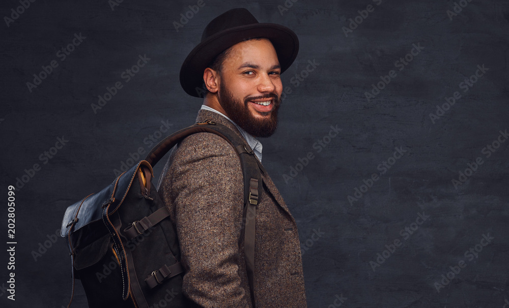 Handsome Afro-American traveler in a brown jacket and hat with the backpack, stands in a studio. Handsome Afro-American traveler in a brown jacket and hat with the backpack, stands in a studio.