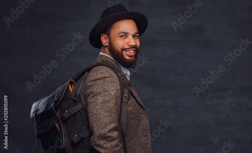 Handsome Afro-American traveler in a brown jacket and hat with the backpack, stands in a studio.