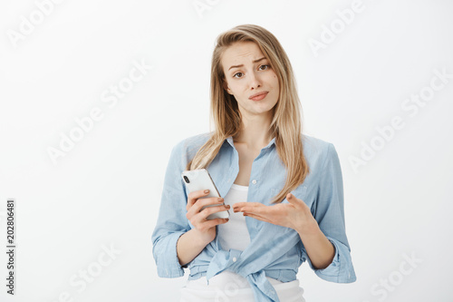 Waist-up shot of annoyed unimpressed attractive european girlfriend with fair hair, smirking, holding smartphone and pointing at device with palm, being shocked with stupid message of ex-boyfriend