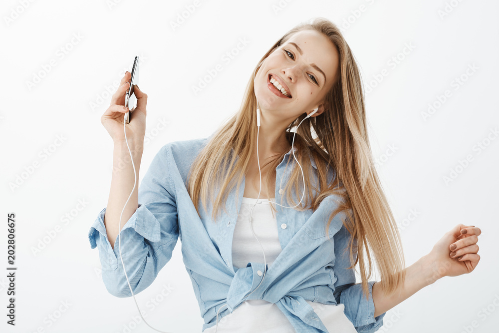 Waist-up shot of outgoing friendly charming woman with fair hair, tilting head while holding smartphone, smiling broadly and listening music in earphones, standing pleased and joyful over gray wall Waist-up shot of outgoing friendly charming woman with fair hair, tilting head while holding smartphone, smiling broadly and listening music in earphones, standing pleased and joyful over gray wall