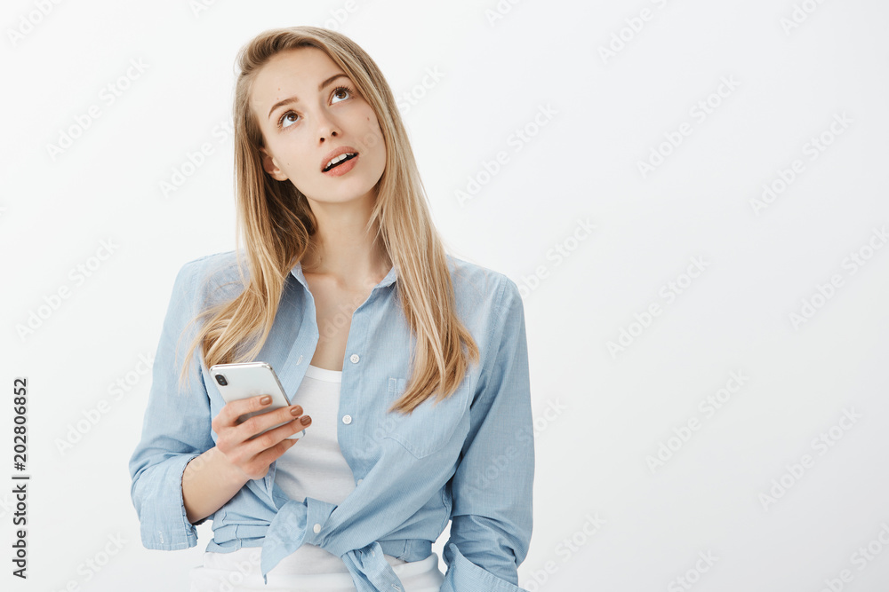 Studio shot of dreamy charming blonde girlfriend in stylish outfit, holdign smartphone and gazing up with interested thoughtful expression, thinking or having something curious in mind Studio shot of dreamy charming blonde girlfriend in stylish outfit, holdign smartphone and gazing up with interested thoughtful expression, thinking or having something curious in mind