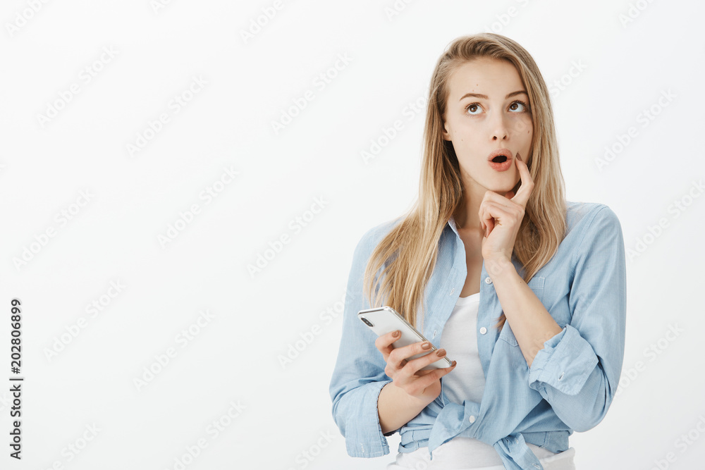 Good-looking dreamy european girlfriend in stylish blue blouse, holding smartphone, biting finger and gazing upwards with curious expression, having idea or thinking up answer on message Good-looking dreamy european girlfriend in stylish blue blouse, holding smartphone, biting finger and gazing upwards with curious expression, having idea or thinking up answer on message