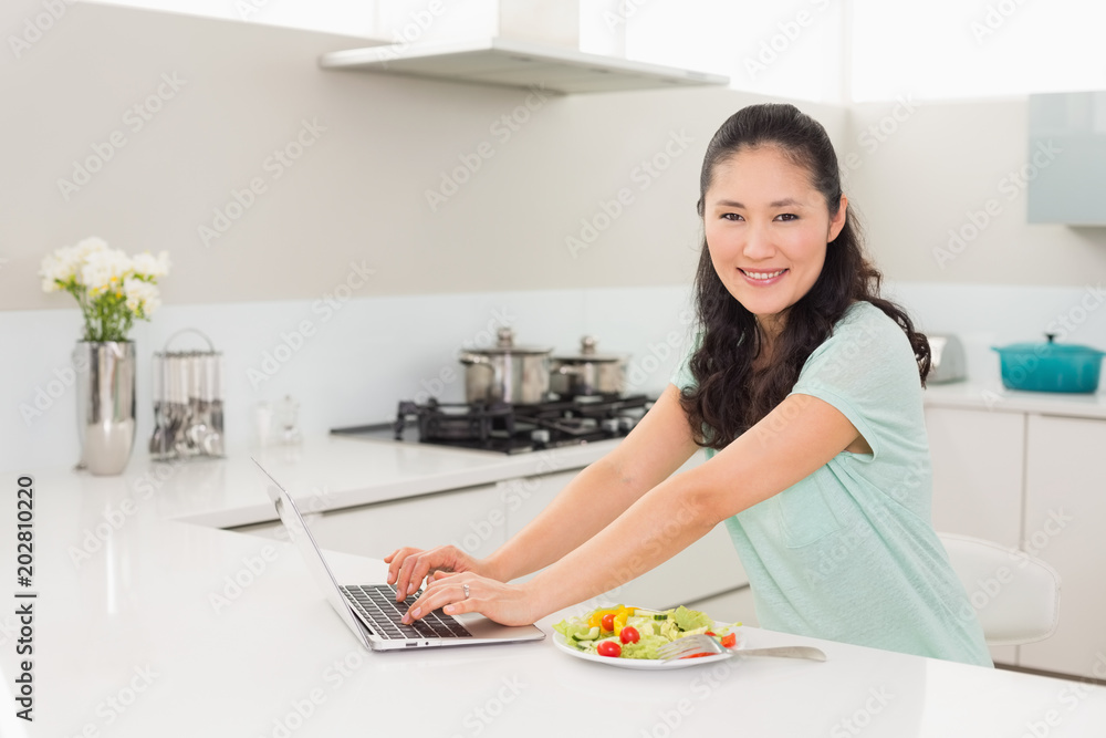 Woman using laptop while having salad in kitchen Woman using laptop while having salad in kitchen