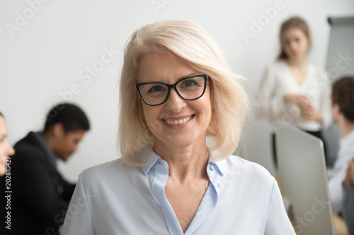 Portrait of smiling senior businesswoman wearing glasses with businesspeople at background, happy older team leader, female aged teacher professor or executive woman boss looking at camera, head shot