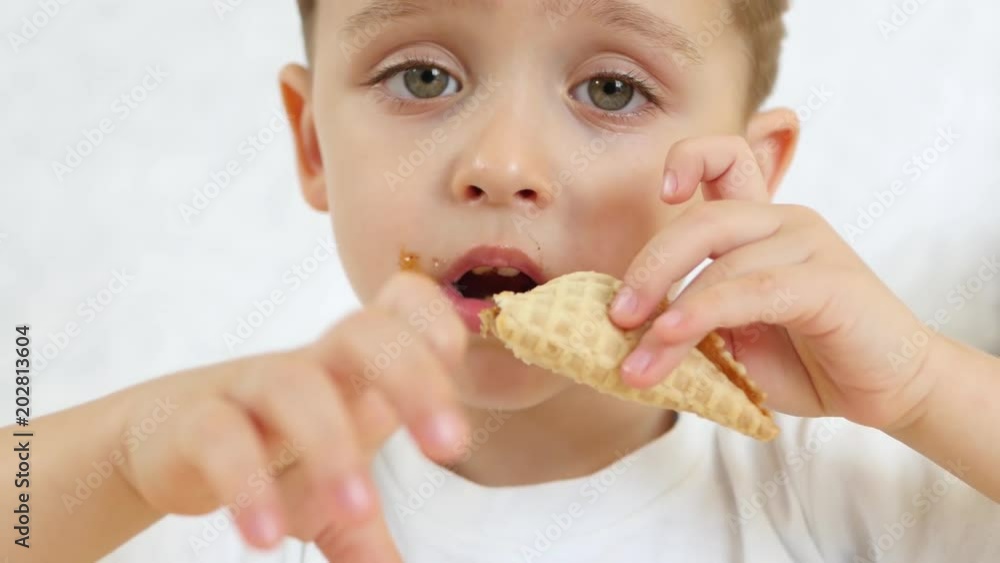 A child is happy to eat a chocolate cone of ice cream in a waffle cone, sitting at a table, on a white background. Focus on ice cream. Close-up. A child is happy to eat a chocolate cone of ice cream in a waffle cone, sitting at a table, on a white background. Focus on ice cream. Close-up.