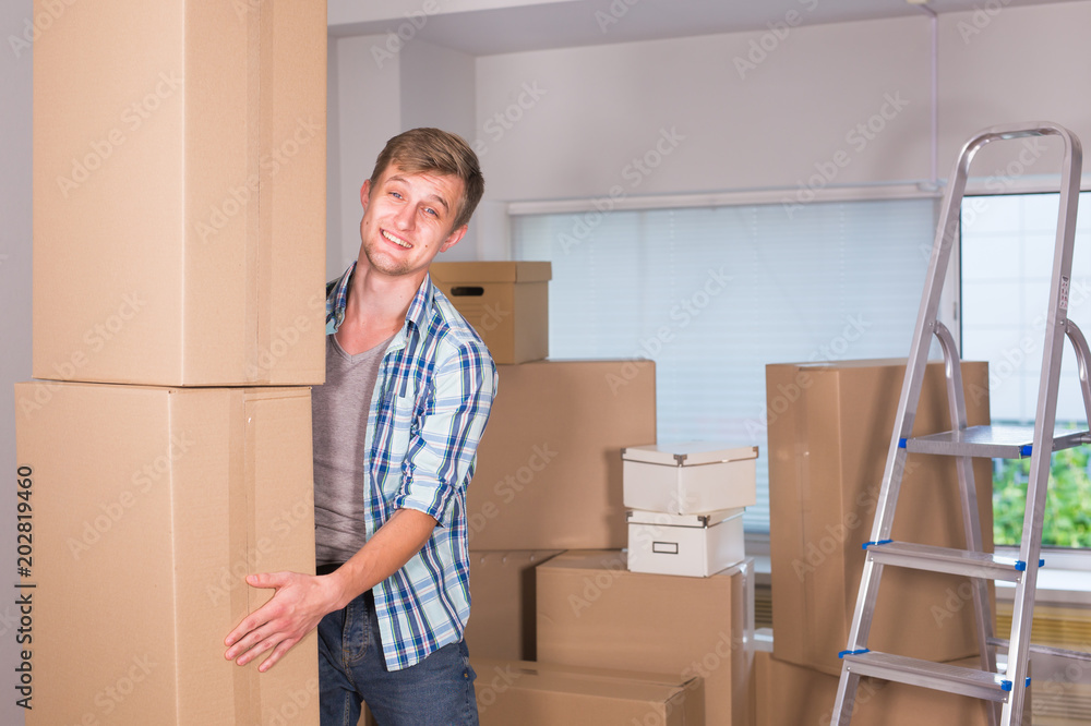 Moving to a new apartment. Cheerful young man holding a cardboard boxes and smiling at camera while other carton boxes laying on background. Moving to a new apartment. Cheerful young man holding a cardboard boxes and smiling at camera while other carton boxes laying on background.