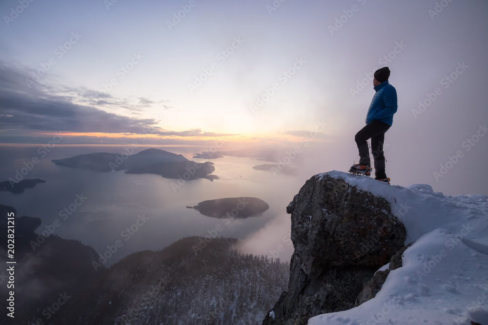 Adventurous man standing on the side of a steep cliff during a beautiful vibrant sunset. Taken on top of St Mark's Peak, North of Vancouver, BC, Canada.